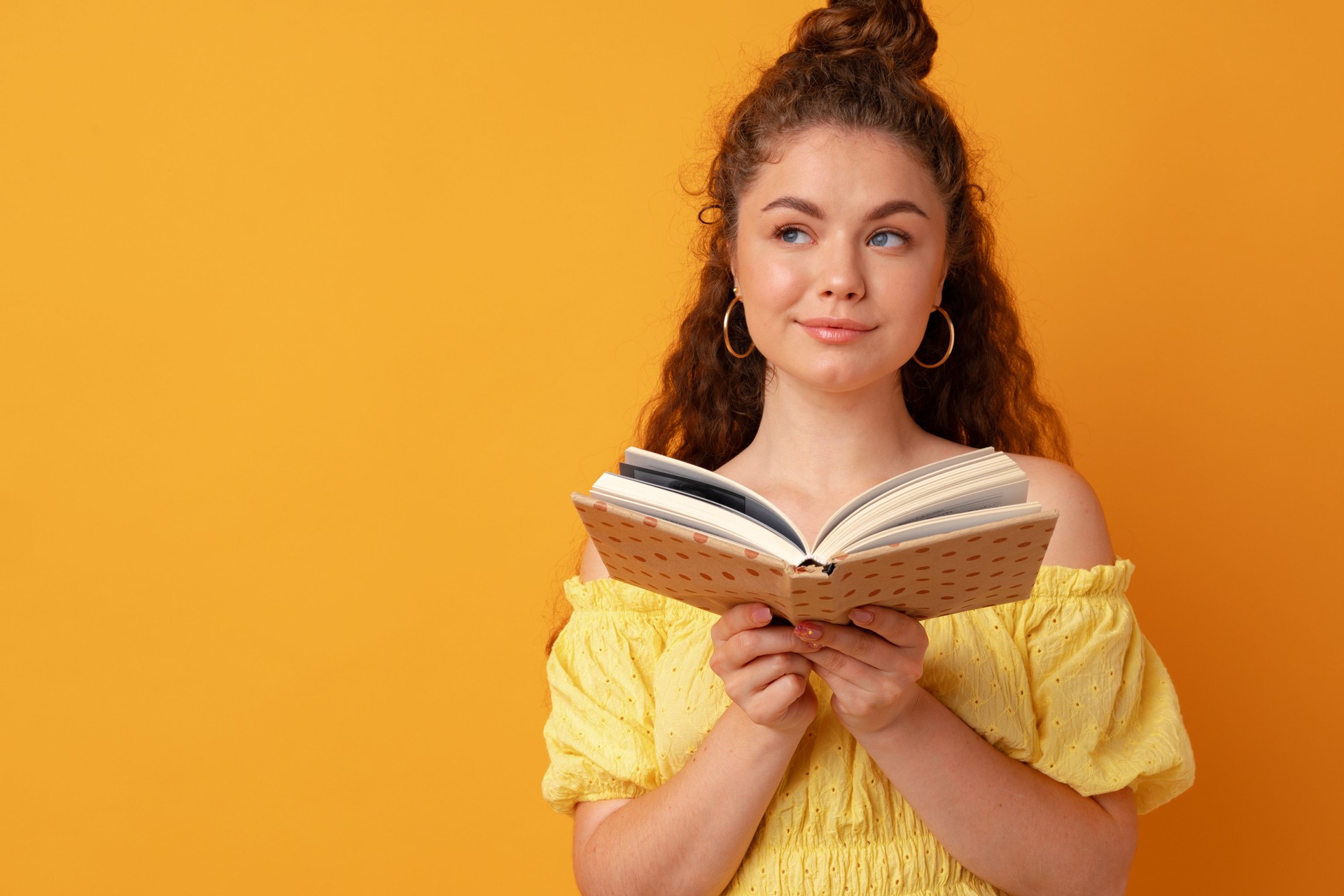 Young curly woman student holding book against yellow background